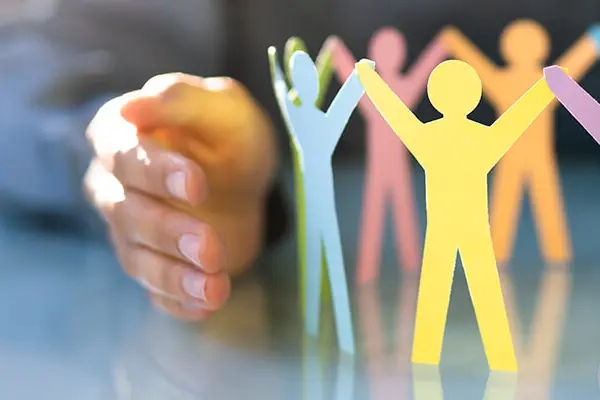 A person is holding up a cut-out paper with small paper figures standing in a row, holding hands. The paper figures appear to form a chain of people holding hands, symbolizing community and togetherness. A person is holding up a cut-out paper with small paper figures standing in a row, holding hands. The paper figures appear to form a chain of people holding hands, symbolizing community and togetherness.